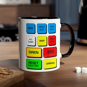 May include: White ceramic mug with a black rim and handle. The mug features a design of a control panel with buttons labelled 360, Front Blue, Rear Blue, H/L Flash, Map, Rear Reds, Siren, 999, Reset Walk Test, and Arrival.