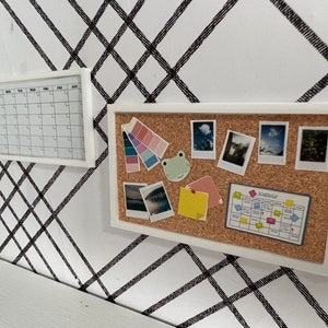May include: A white corkboard with a white frame, featuring a variety of items pinned to it, including photos, a color palette, a frog figurine, sticky notes, and a schedule chart. The corkboard is mounted on a white wall with a black and white criss-cross pattern.
