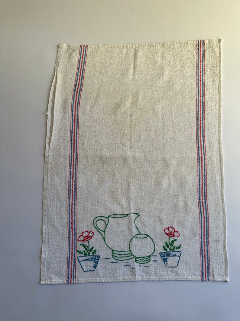 May include: White kitchen towel with red, blue, and green embroidered stripes and a design of a pitcher, a bowl, and two potted flowers with red blooms.