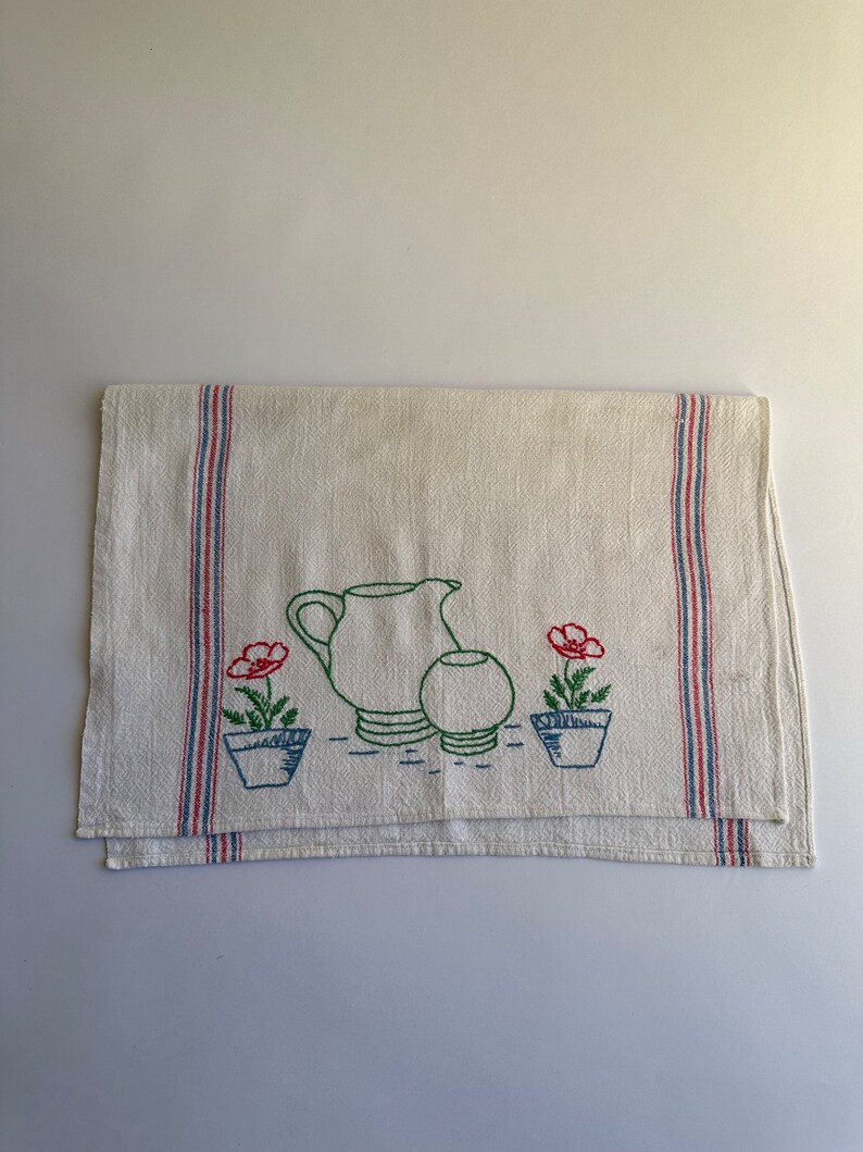 May include: A white tea towel with red and blue stripes along the edges. The towel features an embroidered design of a pitcher, a bowl, and two potted flowers with red blooms.