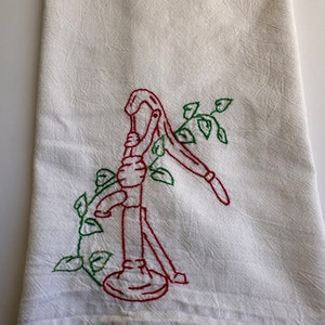 May include: A white tea towel with red and green embroidery of a hand pump with vines.