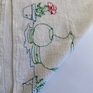 May include: White linen tea towel with embroidered green plants, a red flower, and a blue pitcher. The towel has red and blue stripes along the top and bottom.