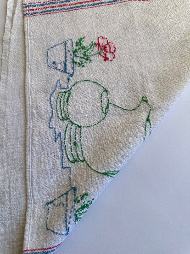 May include: White linen tea towel with embroidered green plants, a red flower, and a blue pitcher. The towel has red and blue stripes along the top and bottom.