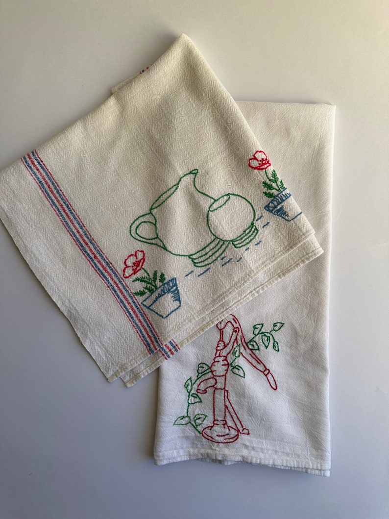 May include: Two white tea towels with embroidered designs. One towel features a pitcher, a bowl, and a flower in a pot. The other towel features a hand pump with vines. Both towels have red, blue, and green embroidery.