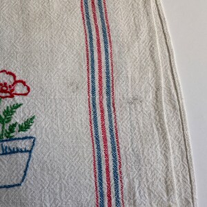 May include: A white linen tea towel with red and blue stripes and a floral embroidery design. The embroidery features a red flower in a blue pot with green leaves.