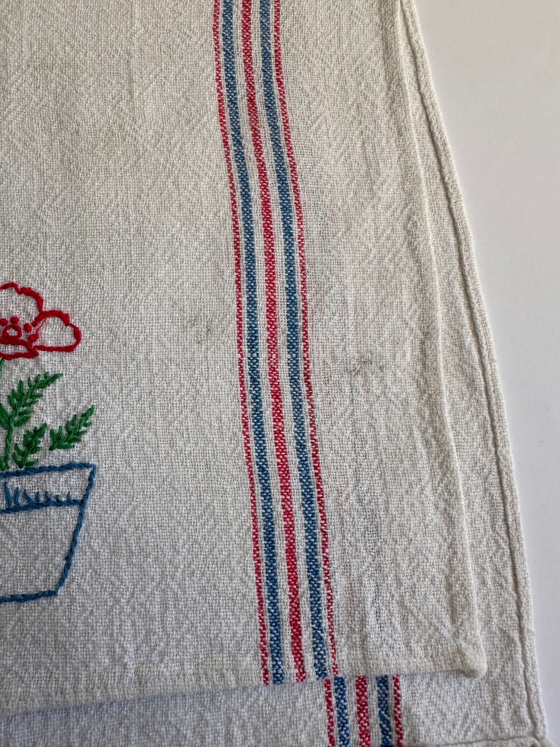 May include: A white linen tea towel with red and blue stripes and a floral embroidery design. The embroidery features a red flower in a blue pot with green leaves.
