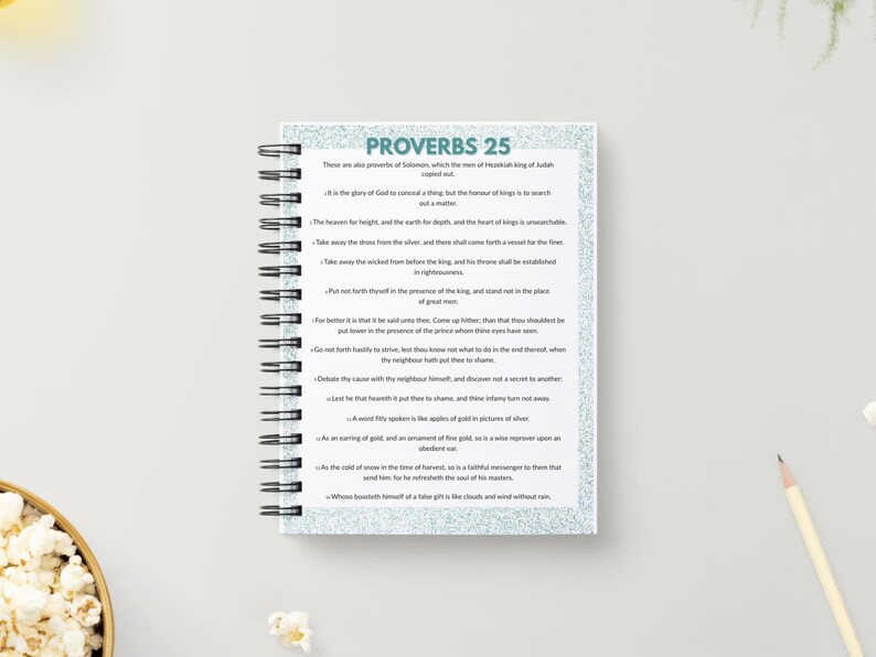 May include: A spiral-bound notebook with the text "PROVERBS 25" in teal. The notebook's pages contain printed text. A pencil and a bowl of popcorn are in the background.