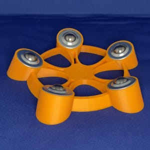 May include: A bright orange circular stand featuring five evenly spaced, raised cylindrical supports. Each support is topped with a smooth, metallic silver ball bearing, creating a star-like configuration with a central open frame design.