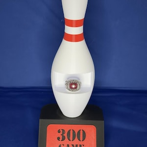May include: A white bowling pin with two red horizontal stripes, featuring a cutout in the center holding a silver ring with a red stone. The pin sits on a black base with a red rectangular plaque displaying the black text 300 GAME.