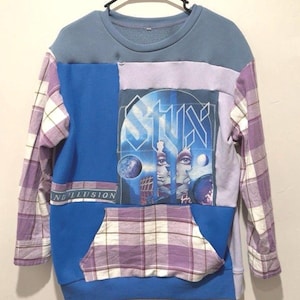 May include: A patchwork sweatshirt with a blue, purple, and plaid design. The front features a graphic of the band STYX with the words "AND ILLUSION". The sleeves are plaid, and the pocket is also plaid.