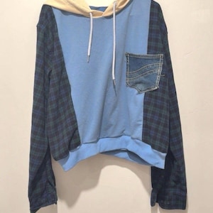 Op de afbeelding: Een lichtblauwe hoodie met een denim zak en geruite mouwen. De capuchon is lichtbeige van kleur en heeft witte trekkoorden. De hoodie hangt aan een zwarte hanger.