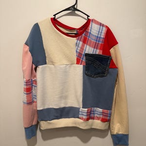 Op de afbeelding: Een patchwork sweatshirt met een spijkerbroekzak. Het heeft panelen van crème, blauwe, roze en rood geruite stof. De top met lange mouwen heeft een rode halslijn en blauwe manchetten, hangend aan een zwarte hanger.