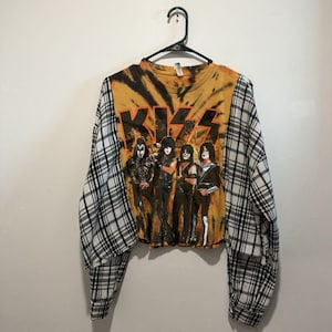 May include: A long-sleeve crew neck shirt featuring a yellow and black tie-dye front panel with a graphic of the band KISS and their logo. The sleeves are made of a white, black, and gray plaid fabric. The shirt has a cropped hemline.