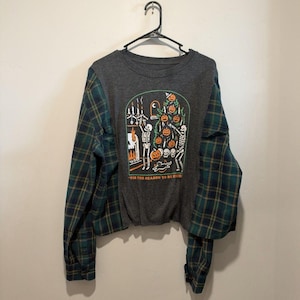 May include: A charcoal gray long-sleeve shirt with green and navy plaid flannel sleeves. The front features a graphic of skeletons decorating a pumpkin tree with the text 'TIS THE SEASON TO BE CREEPY in orange lettering.
