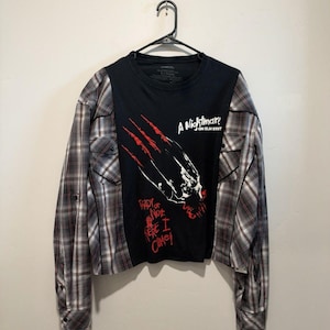 May include: A black long-sleeve shirt with plaid sleeves and a graphic print on the front. The print features a claw mark design, the text A Nightmare on Elm Street, and the phrase Ready or Not Here I Come in red and white lettering.