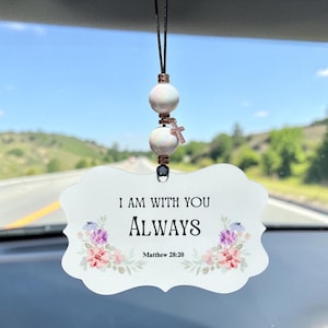 Può includere: Un ornamento da appendere in auto di colore bianco con bordi smerlati, recante il testo I AM WITH YOU ALWAYS e Matthew 28:20 in nero, decorato con fiori ad acquerello rosa, viola e blu, sospeso da un cordino nero con due perline iridescenti e una piccola croce in metallo.