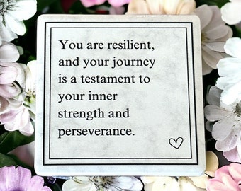 Puede incluir: Una piedra cuadrada blanca con un borde negro y un símbolo de corazón. El texto en la piedra dice "Eres resiliente, y tu viaje es un testimonio de tu fuerza interior y perseverancia."