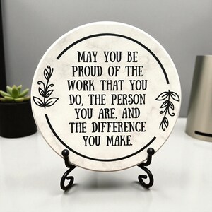 May include: A round, white ceramic coaster with a black border and text that reads "MAY YOU BE PROUD OF THE WORK THAT YOU DO, THE PERSON YOU ARE, AND THE DIFFERENCE YOU MAKE." The coaster is displayed on a black metal stand.