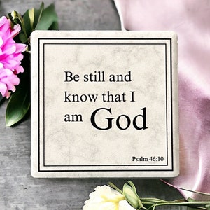 May include: A white square stone coaster with black text that reads "Be still and know that I am God" and "Psalm 46:10".
