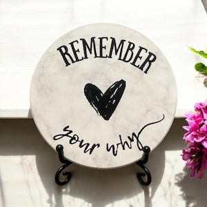 May include: A round white ceramic plate with a black heart and the text "Remember your why". The plate is on a black metal stand.