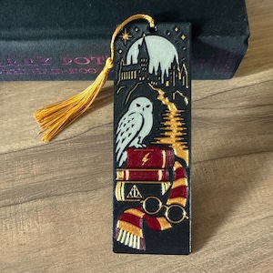 May include: A rectangular black bookmark featuring a gold tassel and an engraved design of a white owl perched on a stack of books. The illustration includes a castle silhouette against a full moon, a lightning bolt, glasses, and a striped scarf.