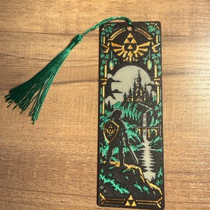May include: A rectangular bookmark featuring a black background with a green and gold illustration of a warrior standing on a cliff overlooking a castle and a river. The design includes a Triforce symbol and a green tassel attached to the top.