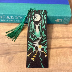 May include: A rectangular black bookmark featuring a green and white graphic of three figures in striped scarves walking toward a glowing stag in a forest under a full moon. A green tassel is attached to the top of the bookmark with a small hole.