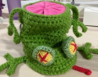 May include: Green crocheted frog toy with yellow eyes and a red tongue. The frog has a pink fabric lining inside its mouth.