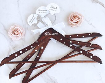 May include: Four wooden hangers with pearl accents and a white satin ribbon. The hangers are personalized with the names "Bride", "Bridesmaid", and "Christine".