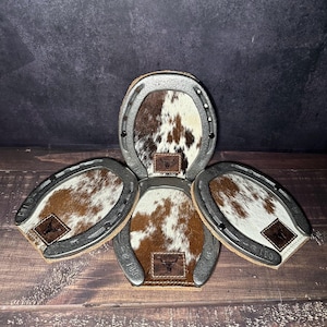 May include: Set of four horseshoe-shaped coasters. Each coaster features a brown and white cowhide center with a leather patch bearing a bull skull logo. The metal horseshoe frames have the number "103" stamped on them.
