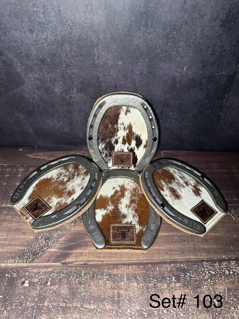 May include: Set of four horseshoe-shaped coasters. Each coaster features a brown and white cowhide center with a leather patch bearing a bull skull logo. The metal horseshoe frames have the number "103" stamped on them.
