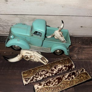 May include: A turquoise vintage-style toy truck with a white bull skull in the bed. Two cowhide and leather decorative items are below the truck, along with another bull skull. The items are on a dark wood surface.