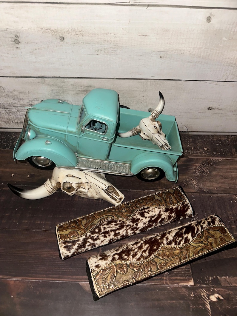 May include: A turquoise vintage-style toy truck with a white bull skull in the bed. Two cowhide and leather decorative items are below the truck, along with another bull skull. The items are on a dark wood surface.