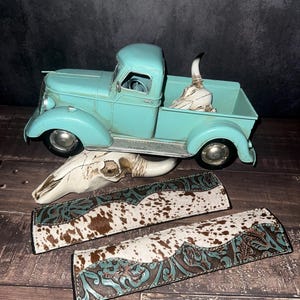 May include: A turquoise vintage pickup truck with a decorative skull in the bed. Two decorative leather straps with cowhide and turquoise floral tooling are in the foreground. A second skull is also present.