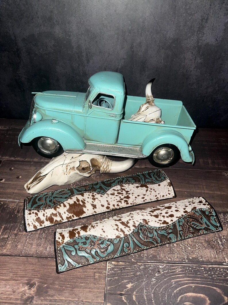 May include: A turquoise vintage pickup truck with a decorative skull in the bed. Two decorative leather straps with cowhide and turquoise floral tooling are in the foreground. A second skull is also present.
