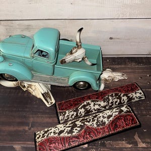 May include: An aqua-coloured vintage truck model with a white steer skull in the bed. Two decorative leather straps with a cowhide pattern and red floral tooling are displayed on a wooden surface. A steer skull is also on the surface.
