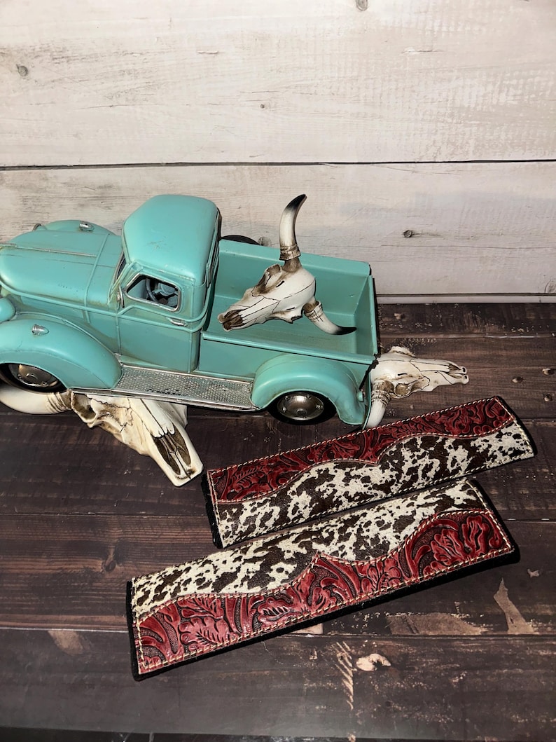 May include: An aqua-coloured vintage truck model with a white steer skull in the bed. Two decorative leather straps with a cowhide pattern and red floral tooling are displayed on a wooden surface. A steer skull is also on the surface.