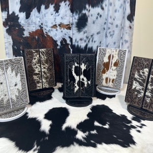May include: Several decorative phone stands with a Western theme. The stands feature a bi-fold design with intricate embossed patterns and cowhide inserts in various colors. Each stand has a circular base and is displayed on a cowhide rug.