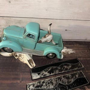 May include: A turquoise vintage truck model with a decorative cow skull and two black and white cowhide leather pieces. The leather pieces have a decorative floral pattern and white stitching.