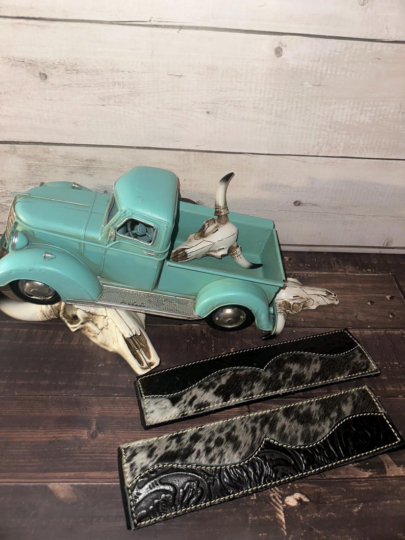 May include: A turquoise vintage truck model with a decorative cow skull and two black and white cowhide leather pieces. The leather pieces have a decorative floral pattern and white stitching.