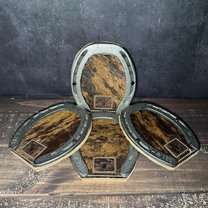 May include: A set of four horseshoe-shaped coasters. Each coaster features a brown and tan cowhide center with a small leather patch and a metal horseshoe frame. The text "Set # 104" is visible at the bottom.