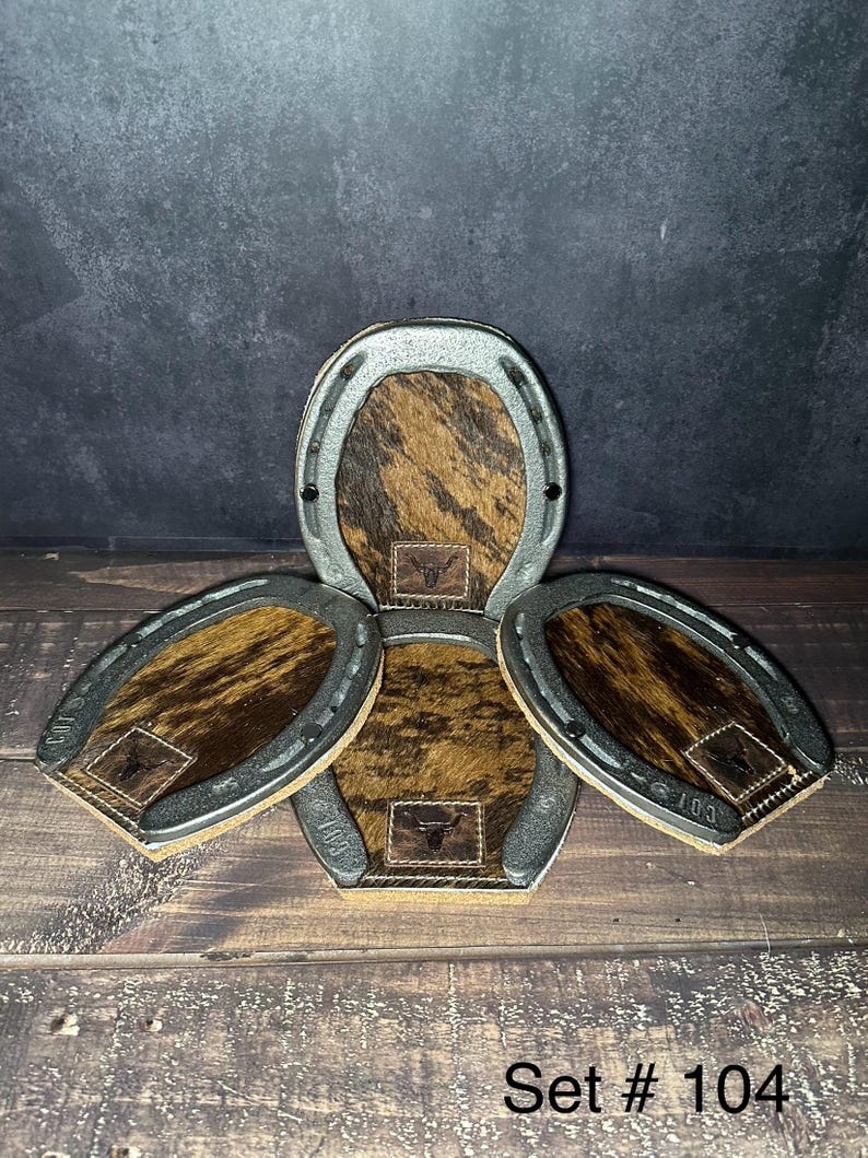 May include: A set of four horseshoe-shaped coasters. Each coaster features a brown and tan cowhide center with a small leather patch and a metal horseshoe frame. The text "Set # 104" is visible at the bottom.