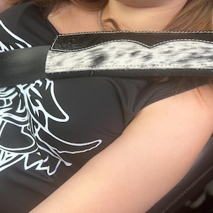 May include: A black and white cowhide seat belt cover with a black border and white stitching. The cover is rectangular and designed to cushion the seat belt. The image also shows a black t-shirt with a white graphic.
