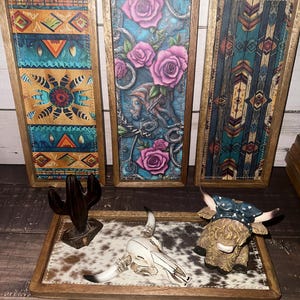 May include: Three rectangular wooden wall art pieces with colorful southwestern designs. A tray with a cowhide pattern holds a cactus, a skull, and a Highland cow figurine.