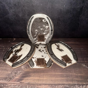 May include: A set of four horseshoe-shaped coasters. Each coaster features a cowhide center with a brown and white pattern, a silver-colored metal rim, and a small leather patch. The text "Set # C106" is visible at the bottom.