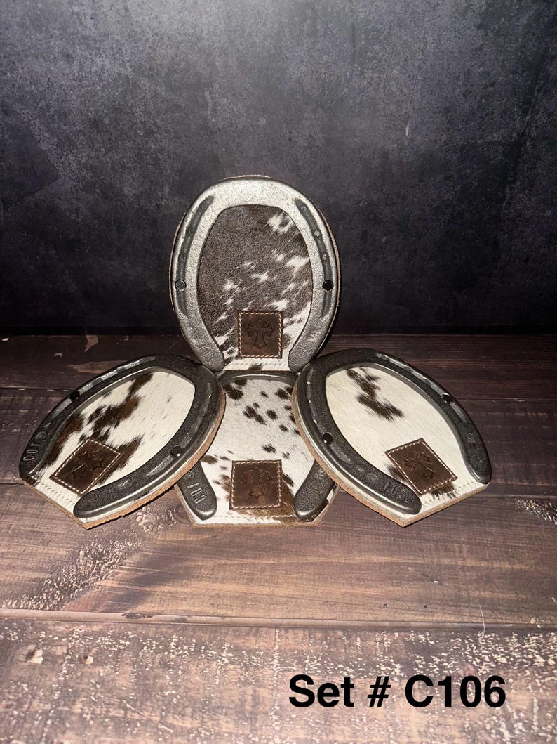 May include: A set of four horseshoe-shaped coasters. Each coaster features a cowhide center with a brown and white pattern, a silver-colored metal rim, and a small leather patch. The text "Set # C106" is visible at the bottom.