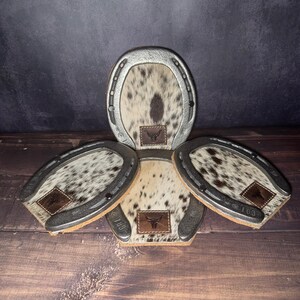 May include: A set of four horseshoe-shaped coasters. Each coaster features a silver-colored metal frame and a cowhide center with brown and white spots. A small leather patch with a bull logo is on each coaster. The text "Set # 100" is at the bottom.
