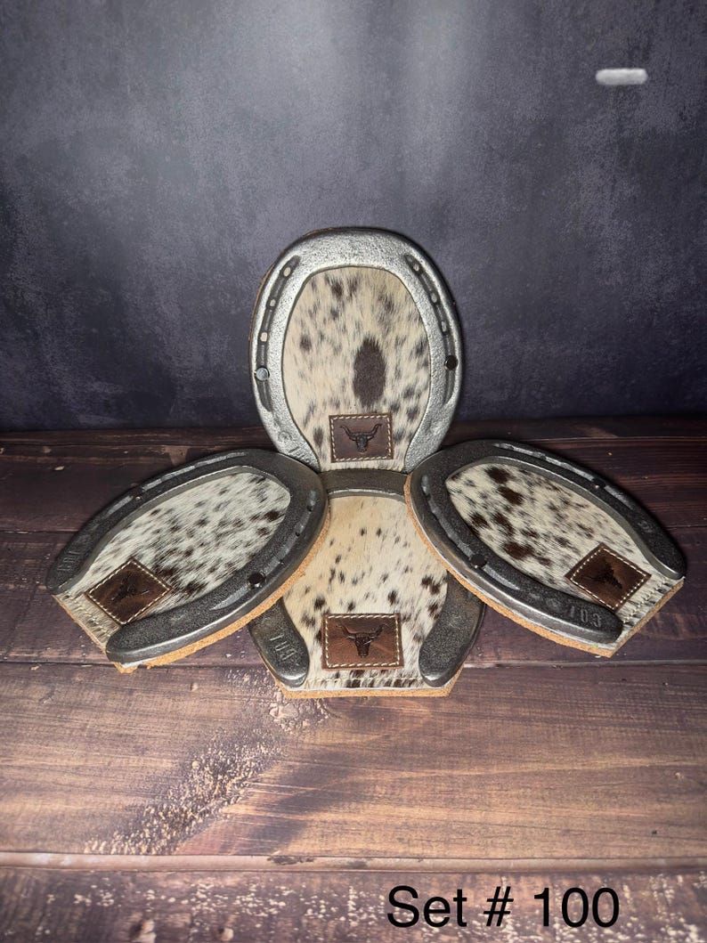 May include: A set of four horseshoe-shaped coasters. Each coaster features a silver-colored metal frame and a cowhide center with brown and white spots. A small leather patch with a bull logo is on each coaster. The text "Set # 100" is at the bottom.