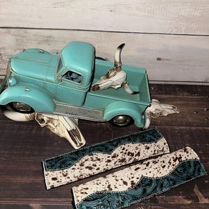 May include: A turquoise vintage truck model with decorative cow skulls and two matching leather accessories with a cowhide pattern and teal accents. The truck is a classic design.