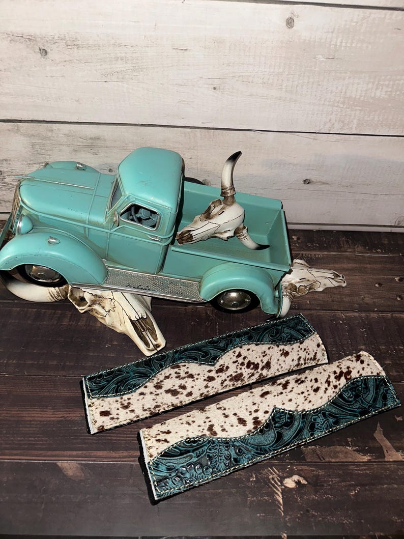 May include: A turquoise vintage truck model with decorative cow skulls and two matching leather accessories with a cowhide pattern and teal accents. The truck is a classic design.
